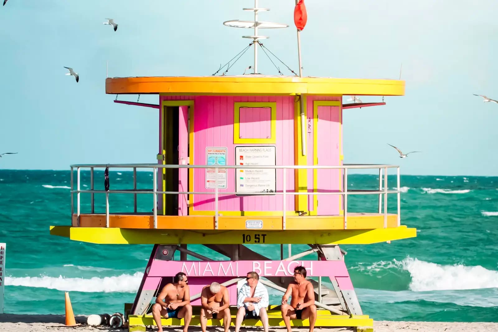 A pink and yellow lifeguard tower on South Beach with people sitting in the shade beneath it