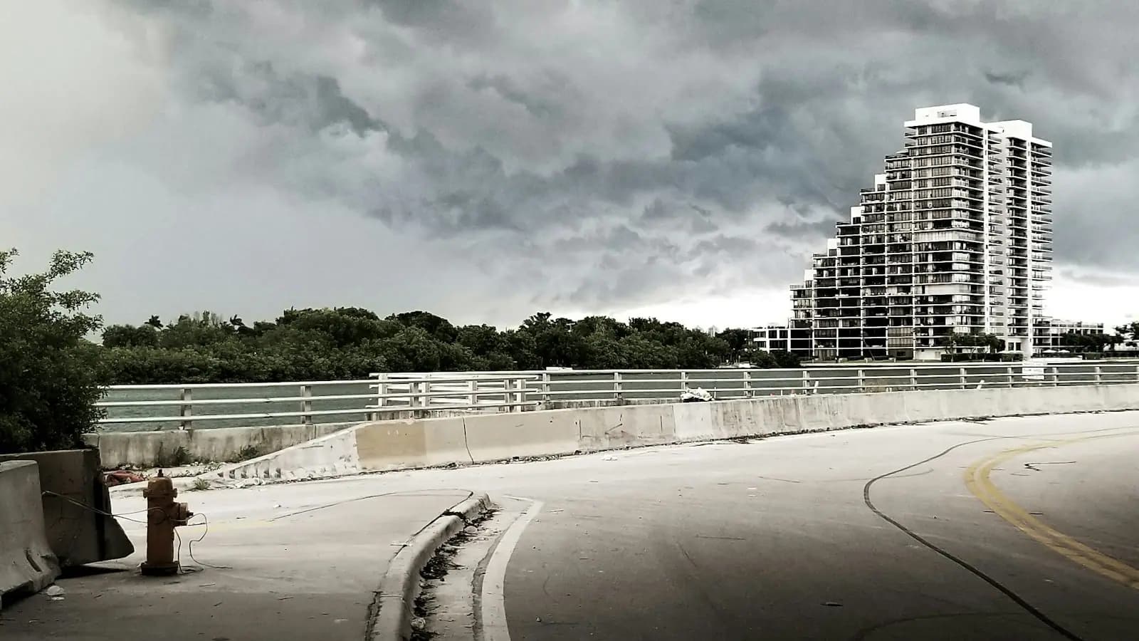 A causeway road with white railings curving towards a Miami high-rise under heavy grey cloud