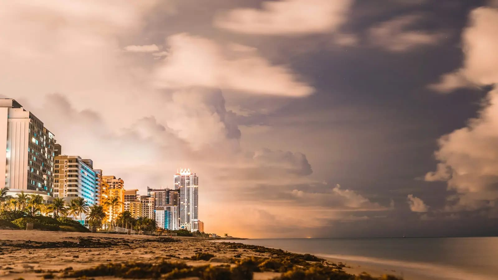 Storm clouds building over the Miami Beach shoreline at dusk with lit hotel towers along the sand